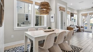 A dining area with a white table and wicker chairs, a hanging light fixture, and large windows with blinds.