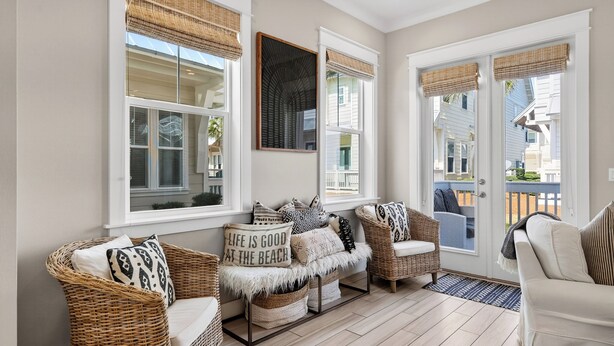 A sunlit room with wicker furniture, a bench with a cushion, and a view of a house through the windows.