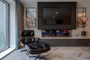 A modern living room with a black leather chair, a large flat-screen TV mounted on the wall, a fireplace with a colorful sofa, and a large window with a view of the city.
