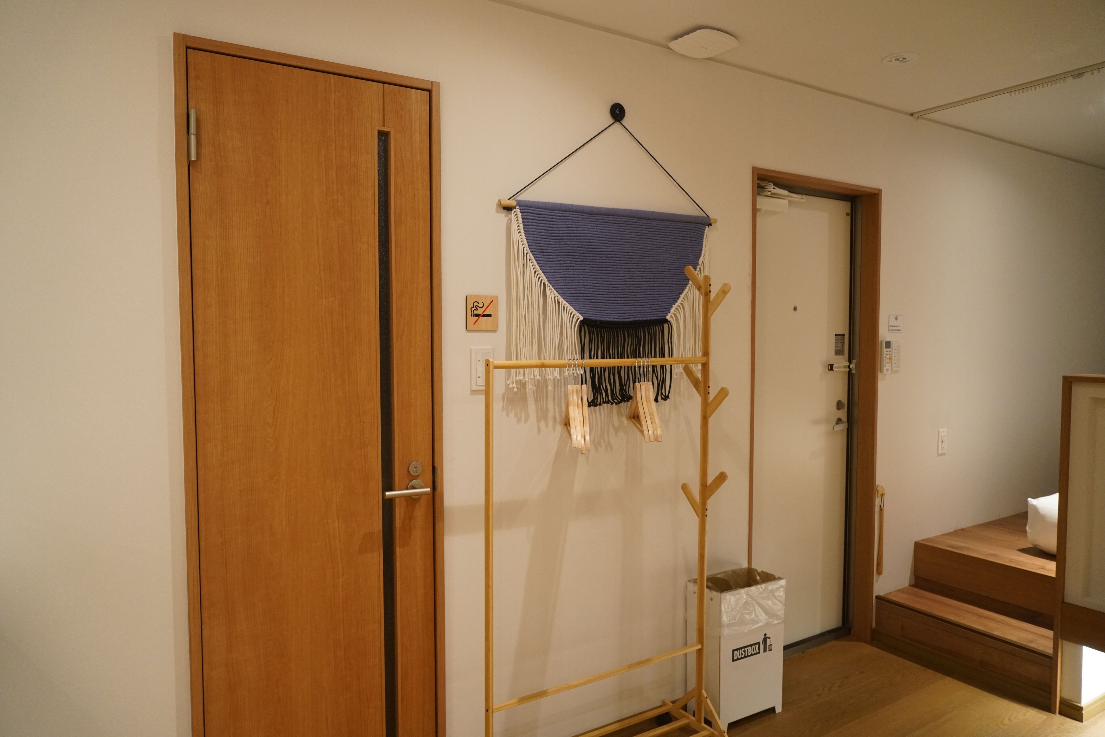 A hallway with wooden doors, a hanging macrame wall hanging, and a small white trash bin labeled "DUSTBOX".