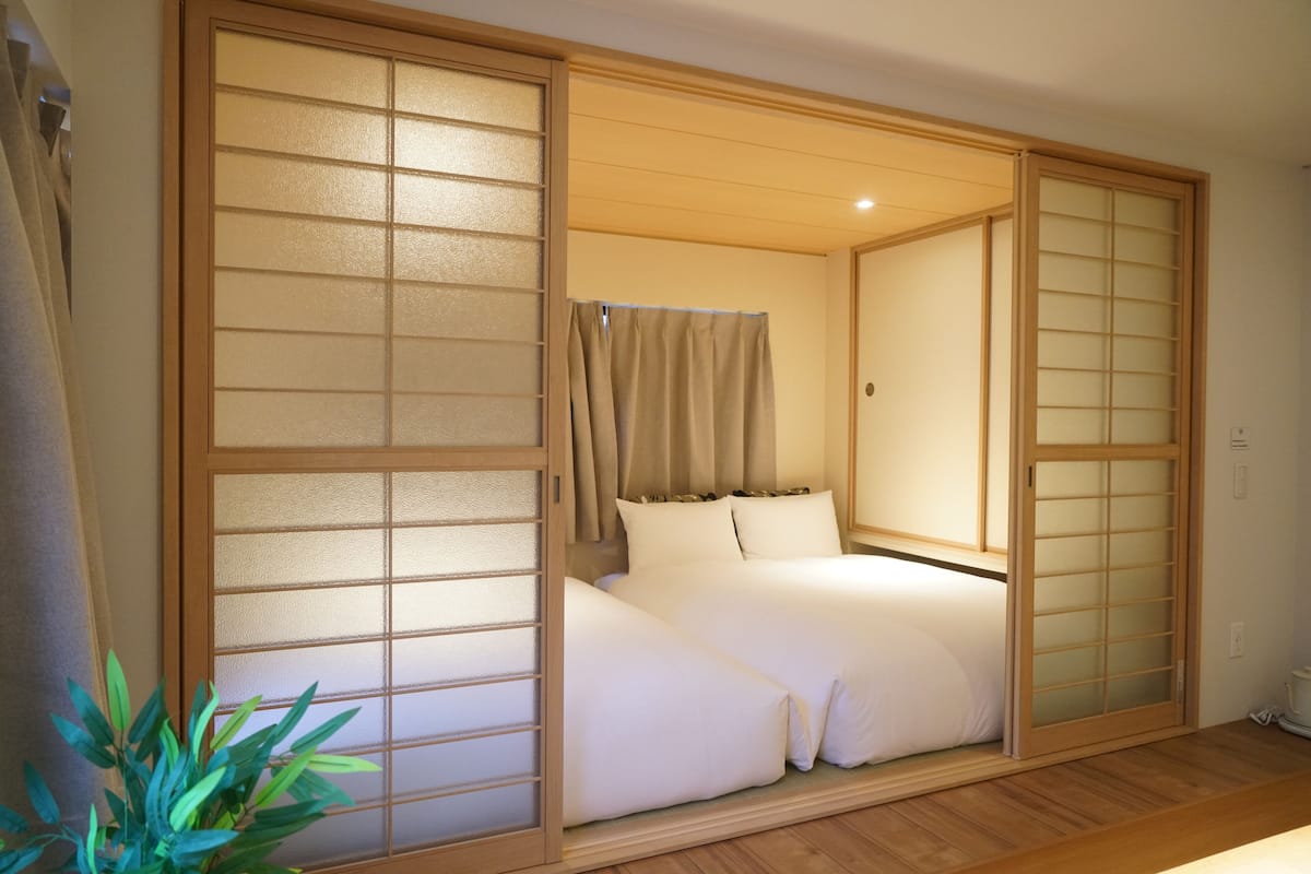 Une pièce de style japonais traditionnel, avec des portes coulissantes, un lit recouvert de draps blancs et une plante en pot.