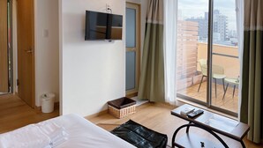 A hotel room with a bed, a TV mounted on the wall, a small table with a remote control, a wicker basket, and a view of the city through a large window with curtains. - softlight inn Asakusa Tokyo (Tokyo)