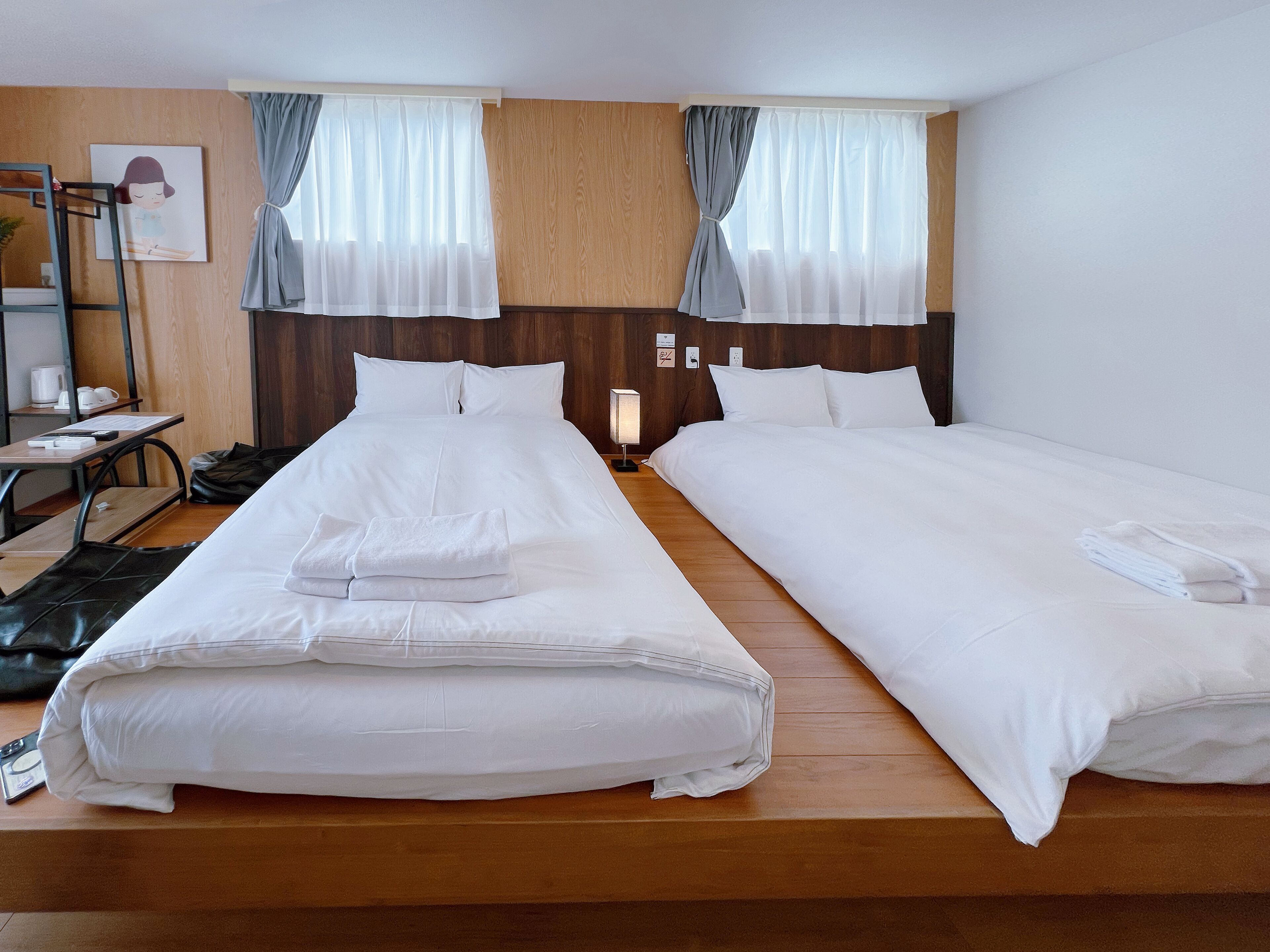 A hotel room with two beds, wooden flooring, a desk with a chair, a small table with a lamp, and a painting on the wall.