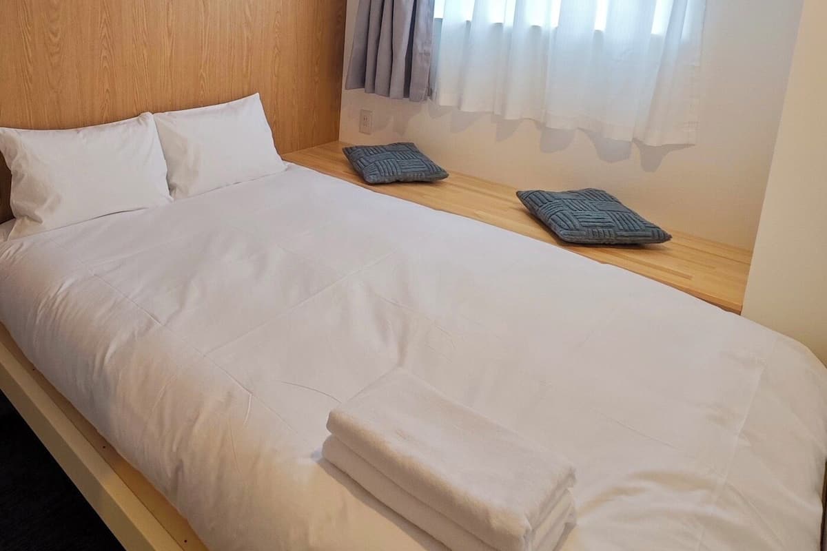 Un lit bien fait, avec des draps blancs et deux oreillers, une tête de lit en bois et une fenêtre ornée de rideaux légers.