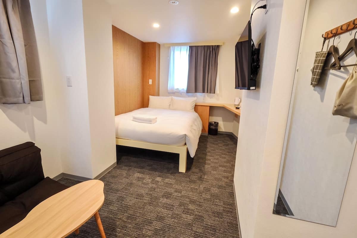 Une chambre d’hôtel de taille réduite, comprenant un lit simple, un petit bureau, un téléviseur fixé au mur et un porte-manteau avec des cintres.