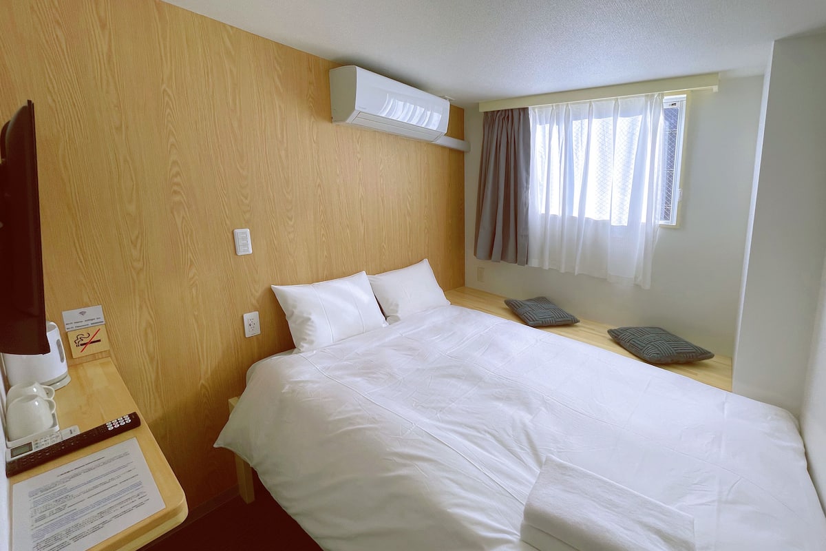 Une chambre d’hôtel avec une tête de lit en bois, un lit avec du linge de lit blanc, une table de chevet avec une télécommande et un menu, et une fenêtre avec des rideaux.