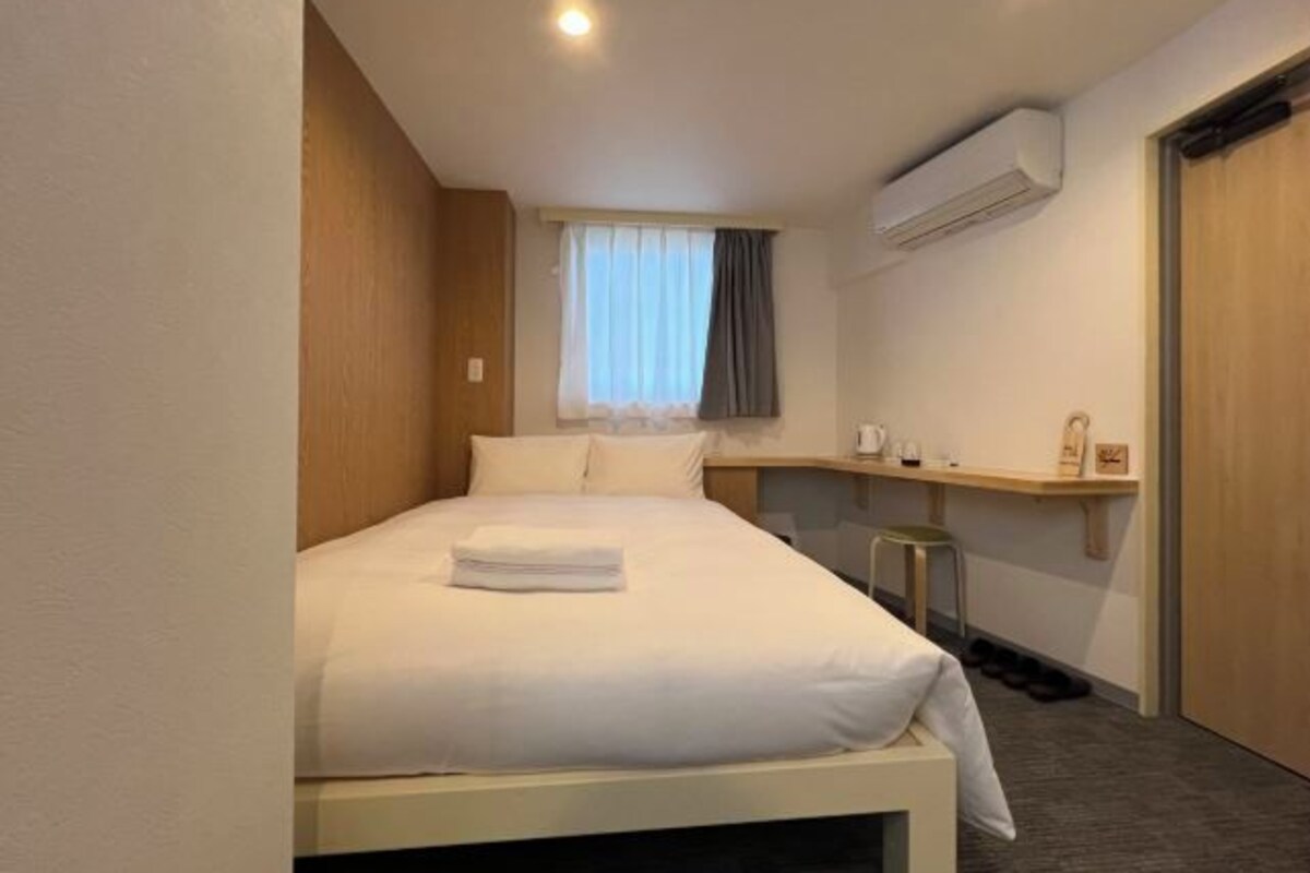 A hotel room with a bed, a small desk, a window with curtains, an air conditioning unit, and a door.