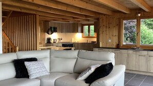 A modern interior with a sofa, wooden beams, a kitchen, and a window with a view of greenery. - Sunny chalet in Veysonnaz with garden (Veysonnaz)