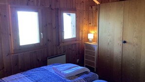 A wooden cabin room with two windows, a bed with a blue patterned bedspread, a nightstand with a lamp, and a wooden wardrobe. - Sunny chalet in Veysonnaz with garden (Veysonnaz)