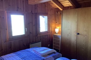 A wooden cabin room with two windows, a bed with a blue patterned bedspread, a nightstand with a lamp, and a wooden wardrobe.