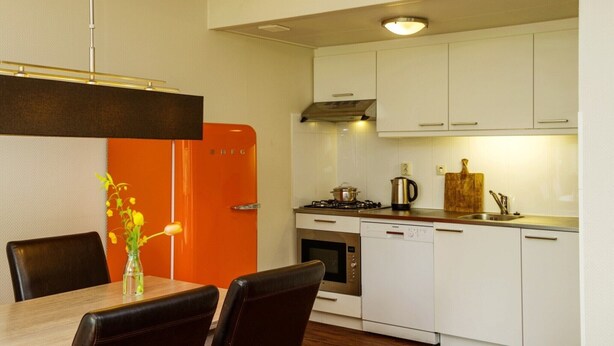 Een moderne keuken met witte kasten, een gasfornuis, een oven en een vaatwasser. Er staat een eettafel met stoelen en een vaas met bloemen.