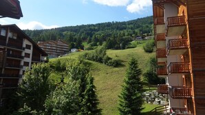 Vista desde la ladera de una colina que muestra un edificio con balcones, rodeado de vegetación y una montaña boscosa al fondo.