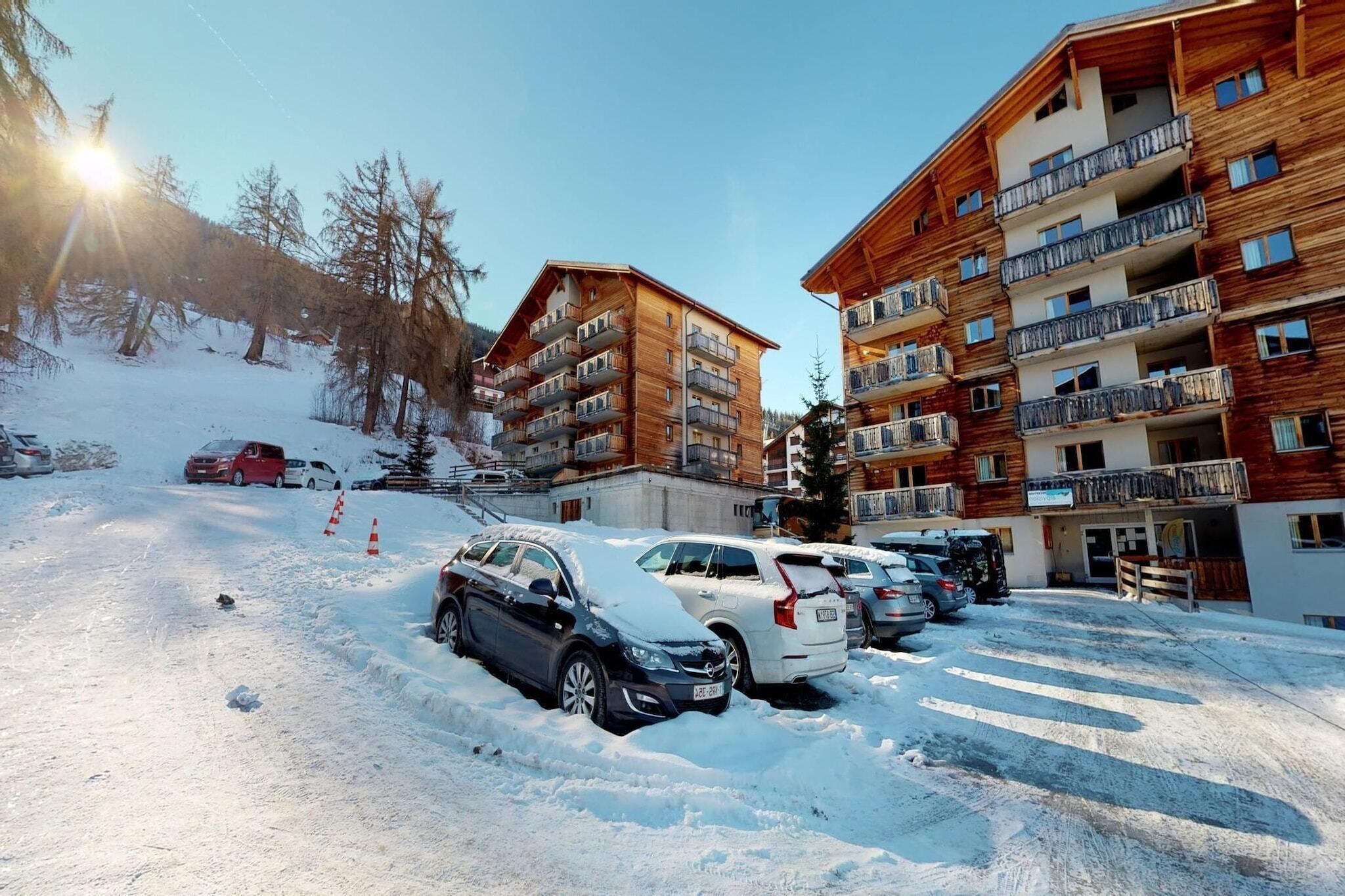 Um estacionamento coberto de neve, com vários carros e um edifício com varandas múltiplas.
