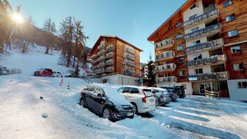 Um estacionamento coberto de neve, com vários carros e um edifício com varandas múltiplas.