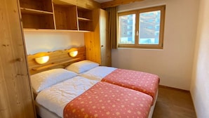 A bedroom with two beds, wooden headboard, built-in shelves, and a window with a view of buildings. - Spacious apartment with view (Haute-Nendaz)
