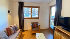 A cozy living room with a sofa, a wooden chair, a television, and a window with a view of buildings. - Apartment near Piste de Tracouet (Haute-Nendaz)