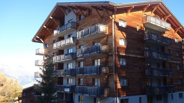 A multi-story wooden building with balconies, likely a hotel or lodge, with a mountainous backdrop. - Welcoming apartment near ski area (Haute-Nendaz)
