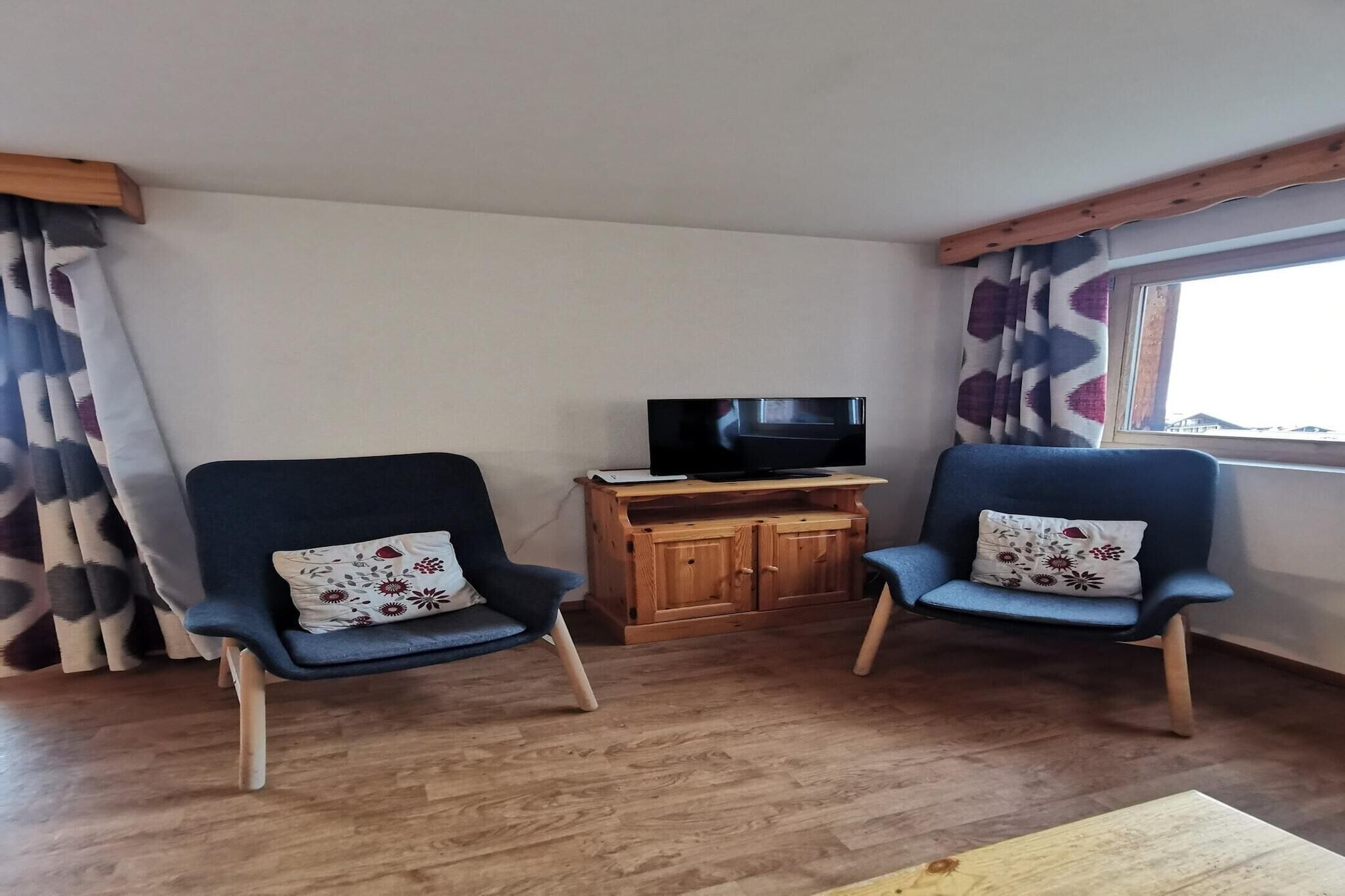 Una habitación con dos sillones azules, un mueble bajo de televisión de madera y una ventana con cortinas.