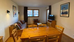 A cozy living room with a wooden dining table, chairs, a sofa, and a television. There are two windows with curtains, and a poster on the wall. - Valley-view apartment with balcony (Haute-Nendaz)