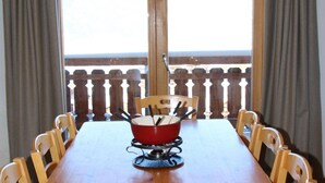 Una mesa de comedor con una olla roja sobre un soporte, rodeada de sillas de madera, con vistas a un balcón al fondo.