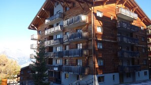 A multi-story wooden building with balconies, likely a hotel or apartment complex, with a view of mountains in the background. - Nice apartment next to cable car (Haute-Nendaz)