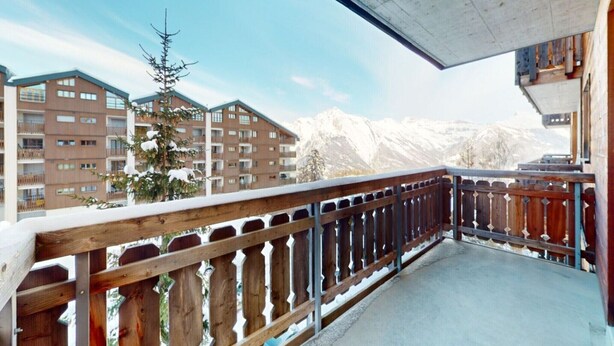 En balkon med trægelænder, udsigt over et bjergrigt område med bygninger i baggrunden.