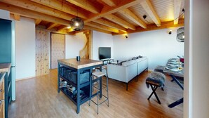 A spacious living area with a wooden ceiling, a sofa, a dining table set for two, and a kitchen area with a bar stool. - Spacious apartment with balcony (Haute-Nendaz)