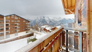A snow-covered mountain view from a wooden balcony with a view of a multi-story building. - Apartment in Nendaz near Cable Car (Haute-Nendaz)