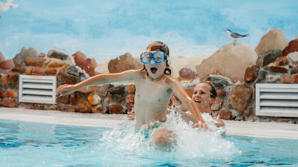 A pool area with a child and an adult in the water, surrounded by a stone wall and a bird perched on a rock.