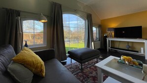 A modern living room with a sofa, ottoman, and a coffee table. There is a TV on a stand and a window with curtains.
