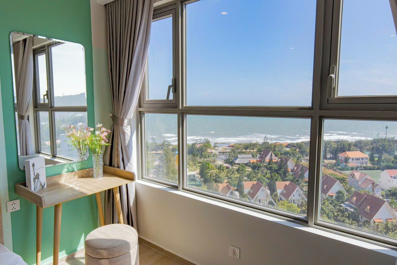 A room with a large window overlooking a coastal area, a wooden table with a vase of flowers, and a mirror on the wall.