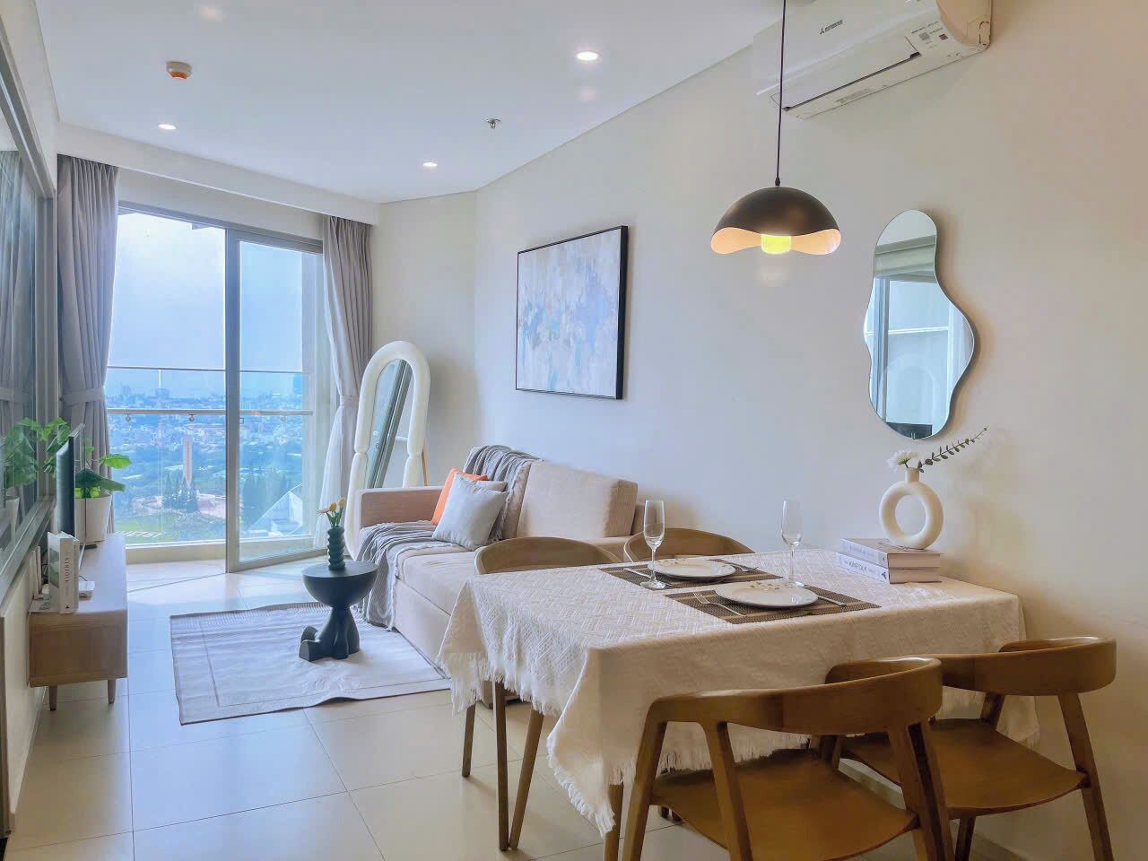 Una habitación de hotel moderna con zona de comedor, sofá, un amplio ventanal con cortinas, ventilador de techo y vistas a la ciudad.