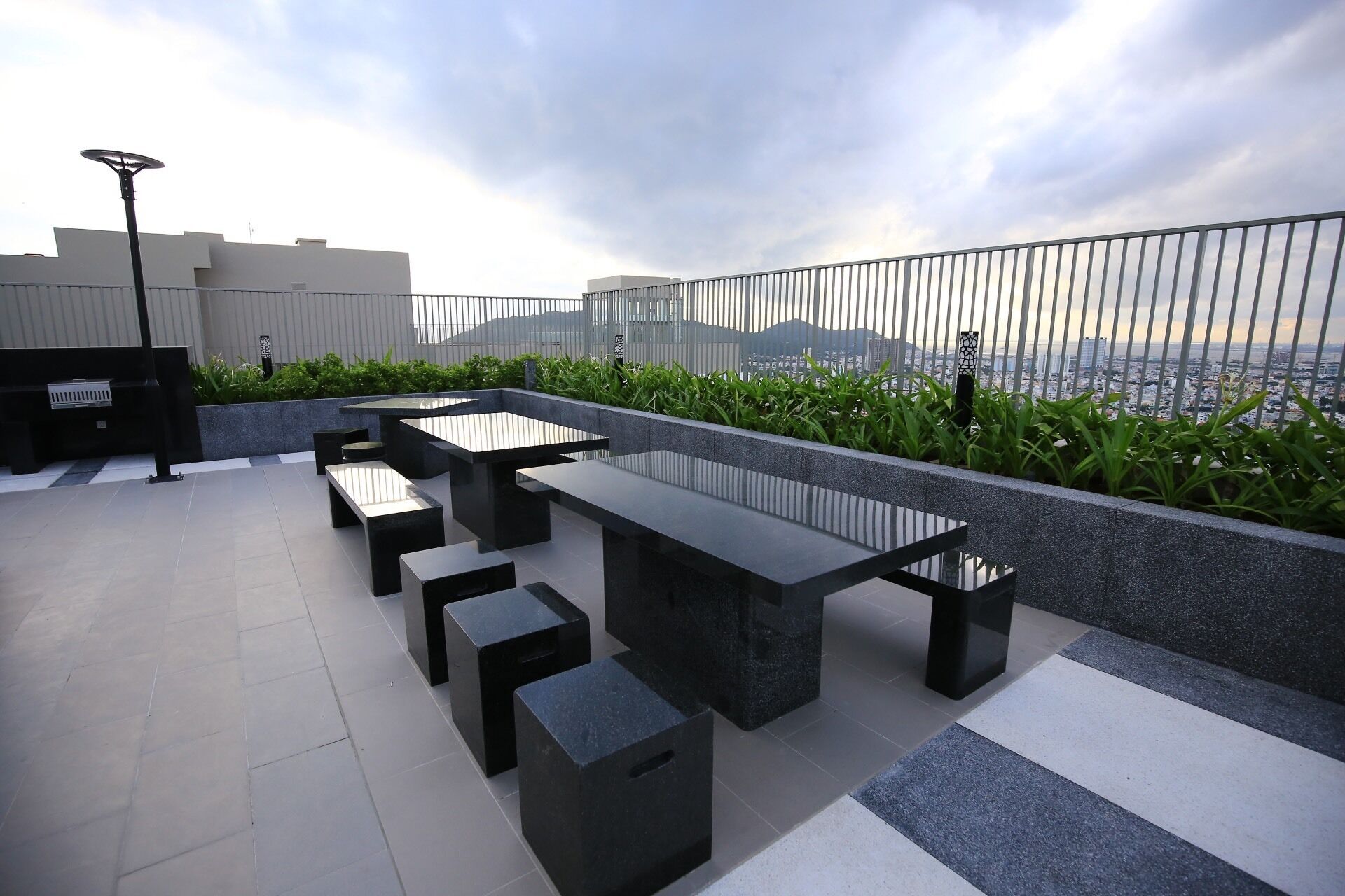 Zona de estar al aire libre con varias mesas y bancos negros, rodeada por una barandilla de metal, con vistas a un paisaje urbano.