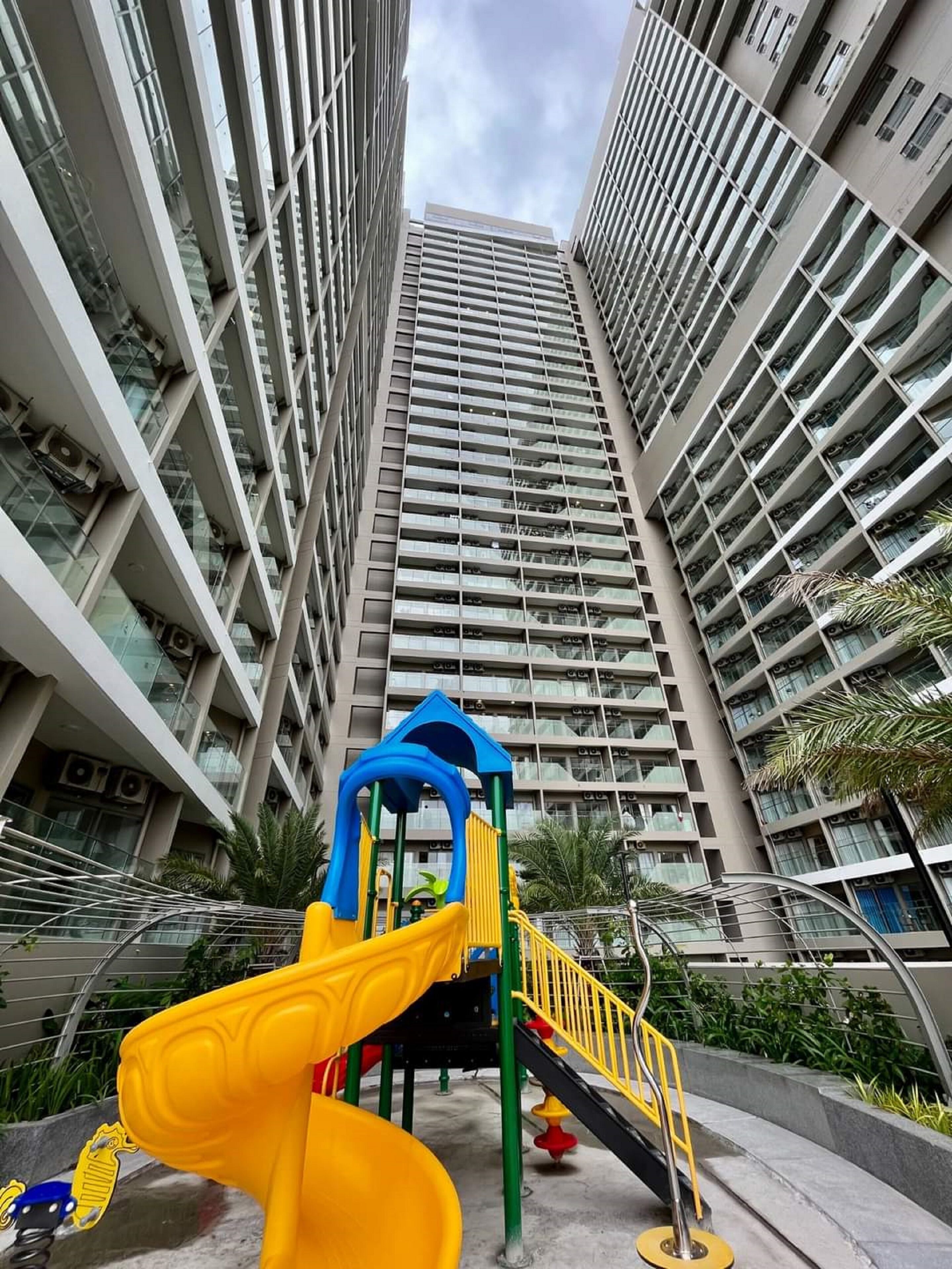 A playground with a yellow slide and a blue climbing structure, surrounded by tall residential buildings.