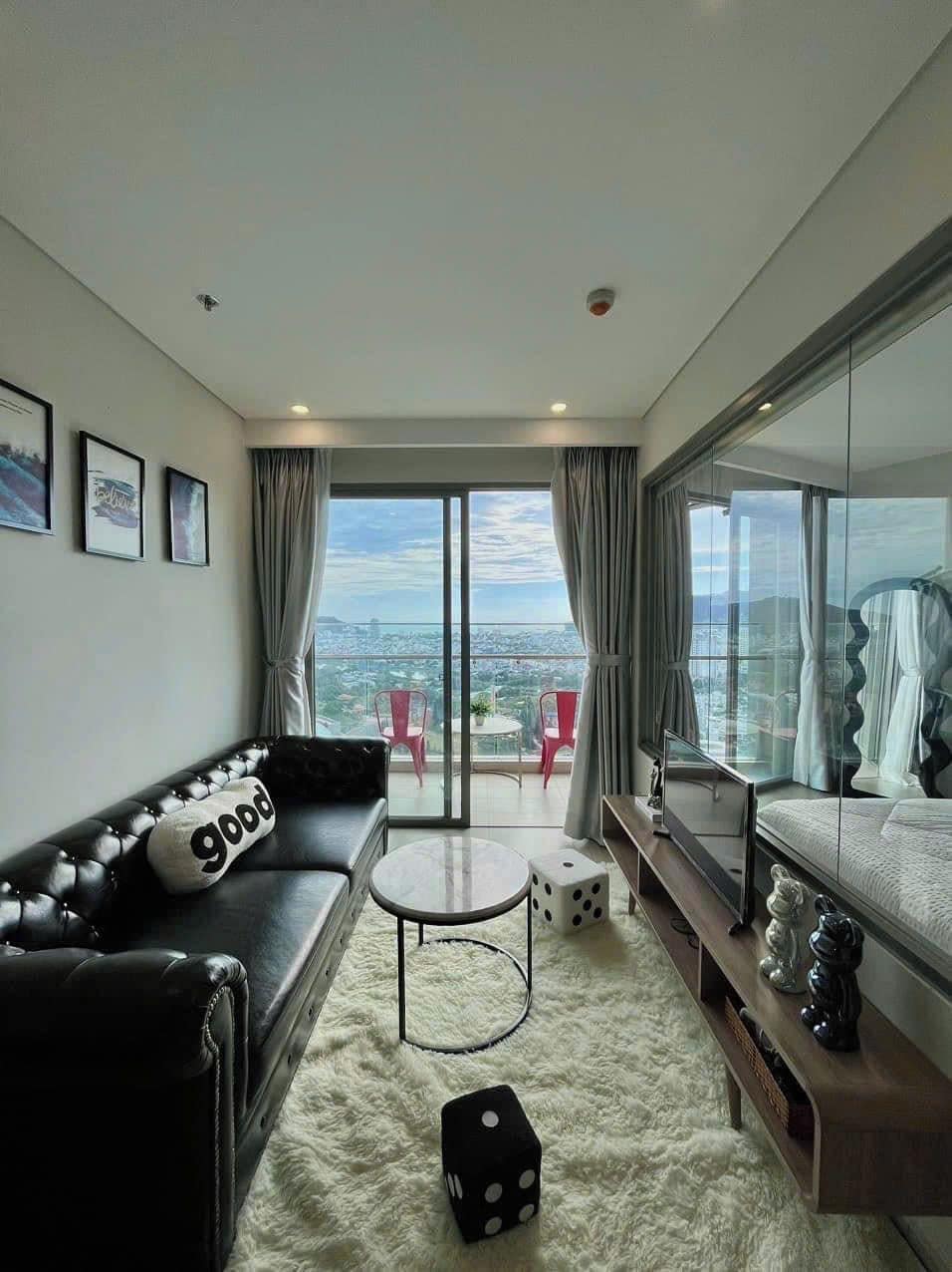 A modern hotel room with a leather sofa, a small round table, a TV, and a balcony with red chairs.
