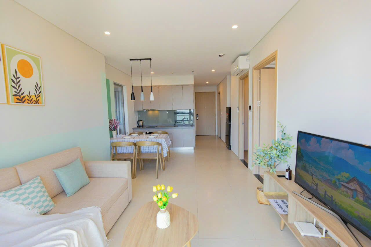 A modern living room with a sofa, a coffee table with flowers, a TV, and a kitchen in the background.