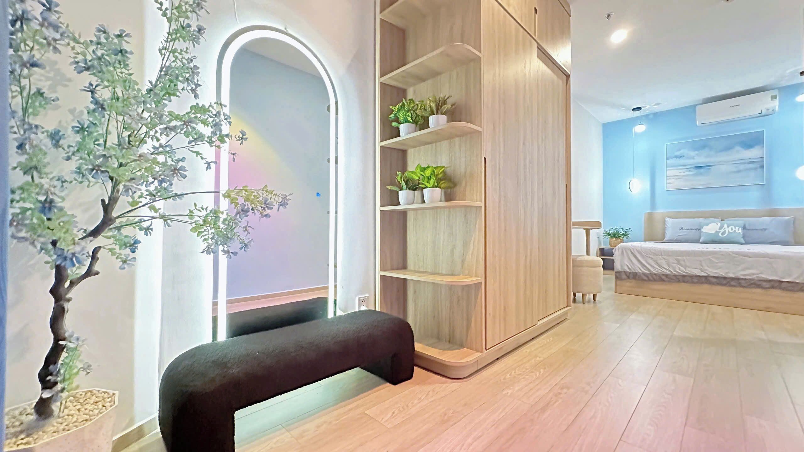 Un dormitorio moderno con un armario de madera, un banco, una cama con almohada azul y un estante con plantas.
