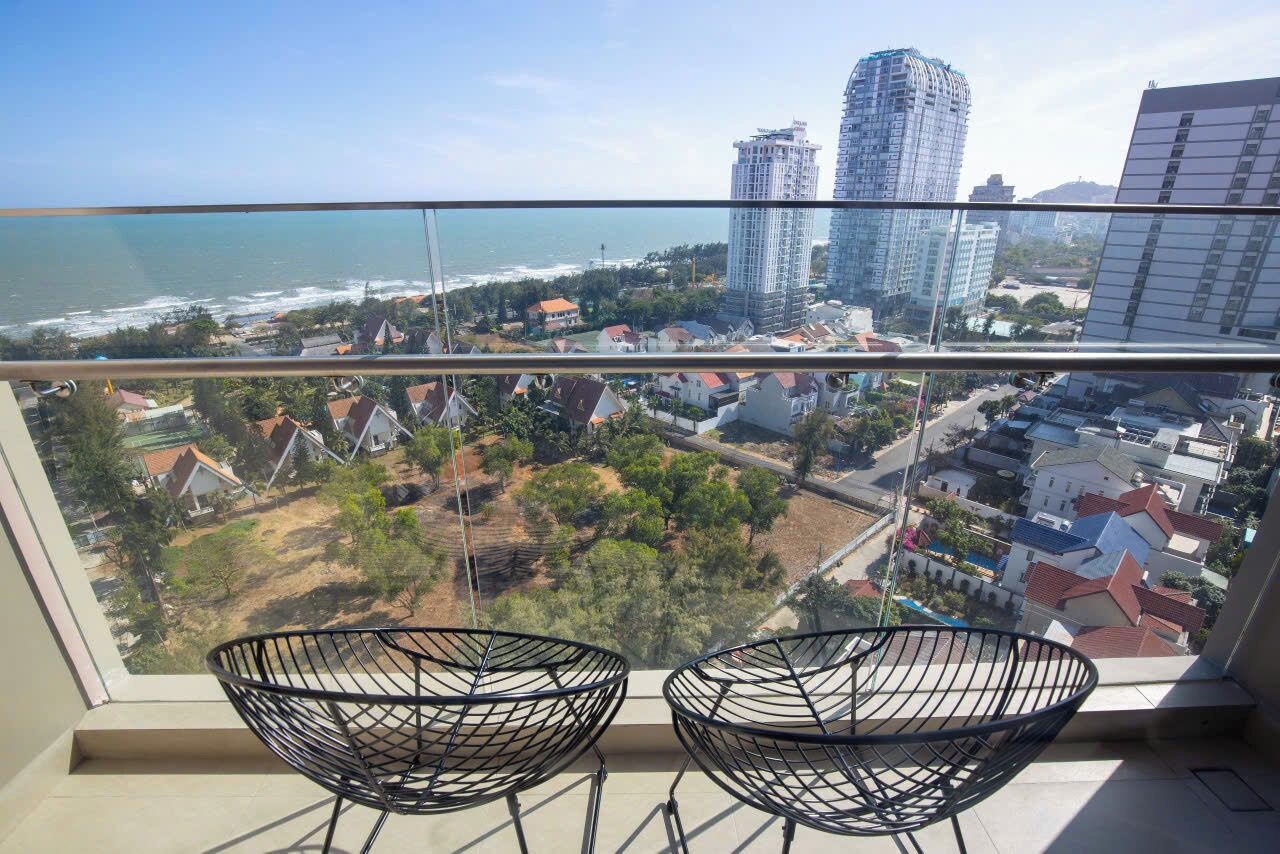 Un balcón con dos sillas de alambre negras, con vista a una ciudad costera con edificios y una playa.