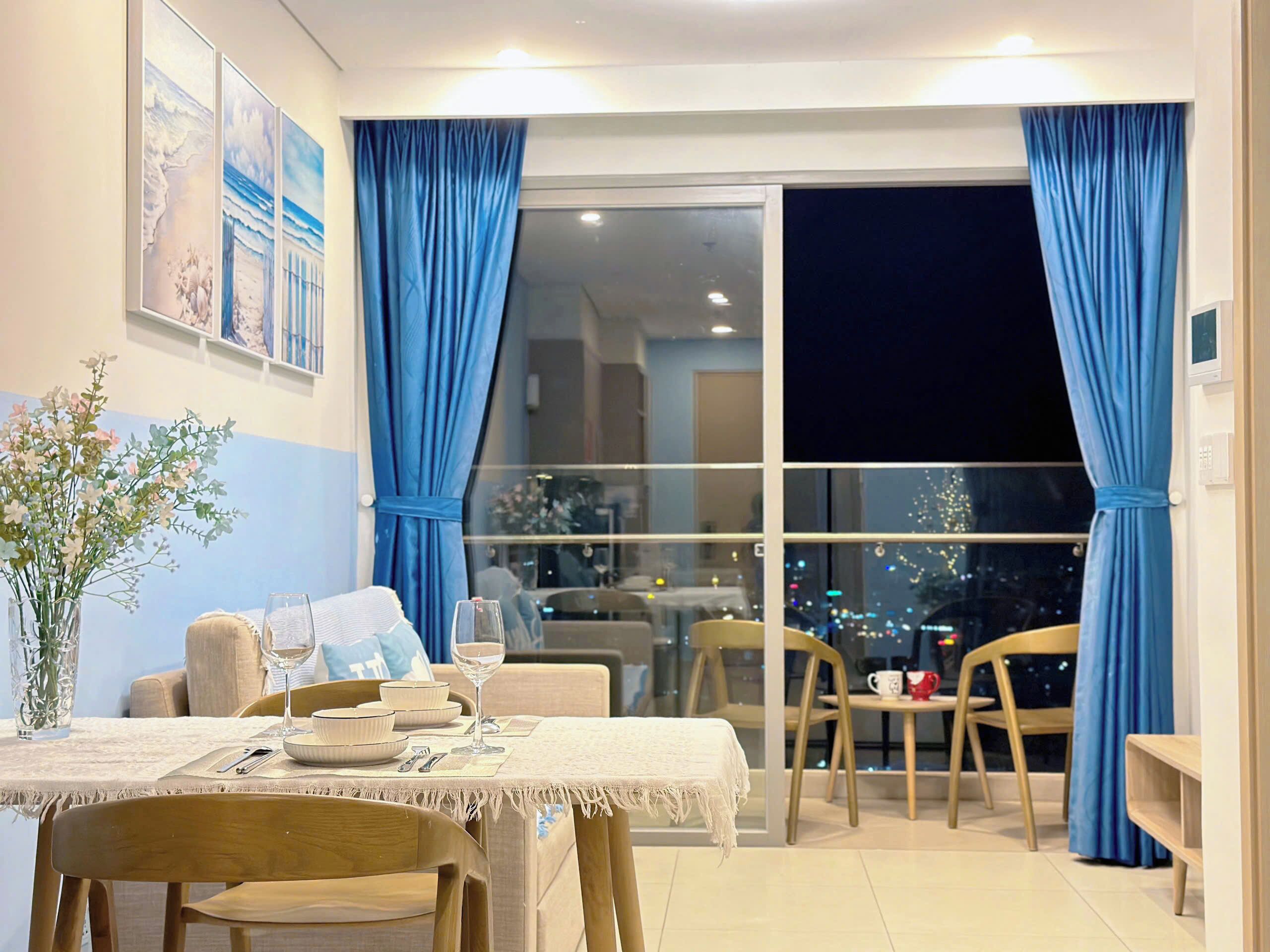 A dining area with a table set for two, a vase with flowers, and a view of the city at night through large windows.
