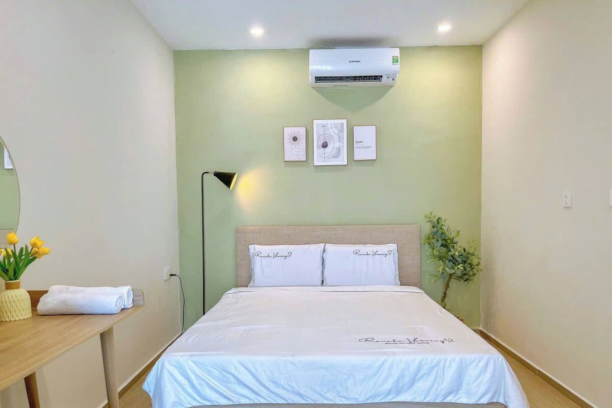 Habitación de hotel ordenada con una cama, una mesita de noche con lámpara, una planta pequeña y un aire acondicionado de pared.