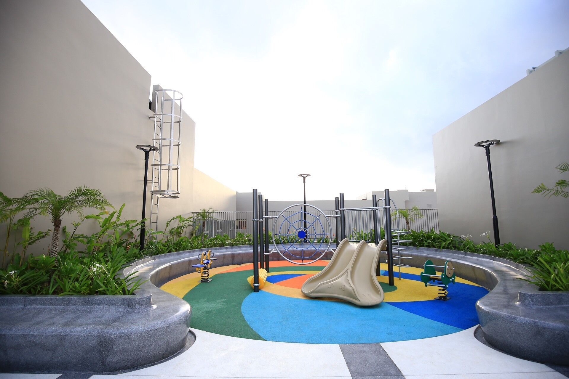A playground with a colorful play area, including slides and swings, surrounded by planters and a clear sky.