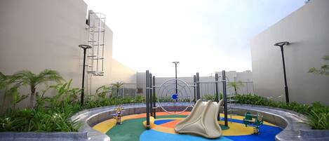 A playground with a colorful play area, including slides and swings, surrounded by planters and a clear sky.