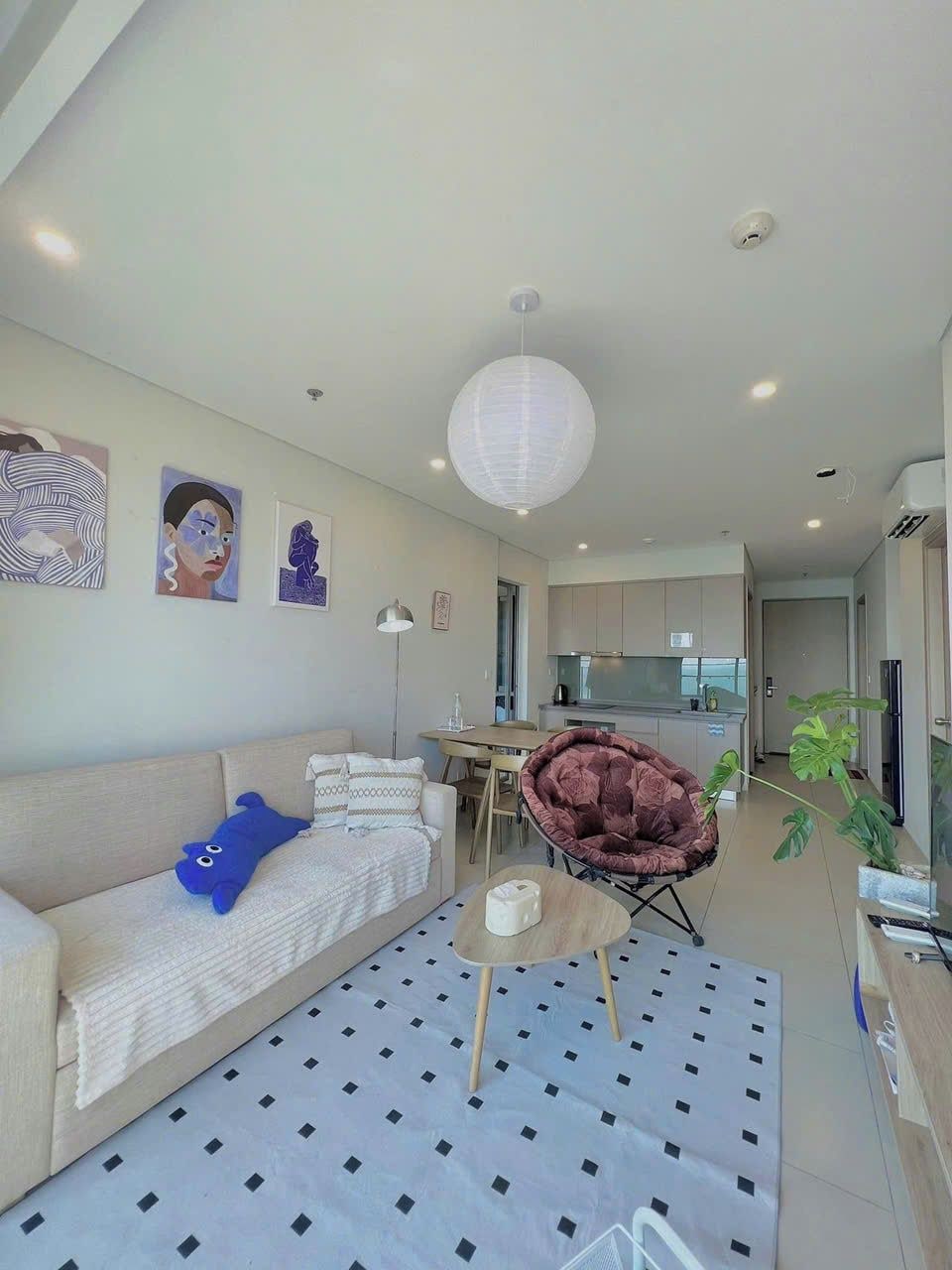 A modern living room with a sofa, a blue plush toy, a patterned rug, a hanging light fixture, and a dining area with a chair and table.