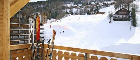 A wooden balcony with ski equipment, a view of a snowy mountain slope, and buildings in the background.