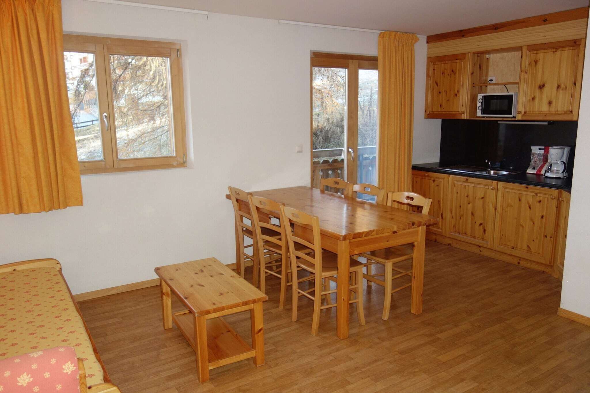 Une pièce de taille modeste comprenant une table à manger en bois, des chaises, un petit banc en bois, un canapé et une kitchenette dotée de meubles de rangement en bois et d’un four à micro-ondes.