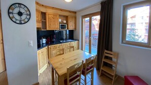Une cuisine compacte avec des meubles en bois, un four à micro-ondes et un évier. On y trouve une table à manger avec des chaises et un banc rouge. Une horloge est fixée au mur, et une fenêtre offre une vue sur l’extérieur.