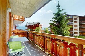 A balcony with a wooden railing, a table with chairs, and a view of a mountain and buildings in the background.