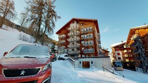 A multi-story wooden building with balconies, a snow-covered road, and a red car parked in front. - Appealing apartment with balcony (Haute-Nendaz)