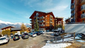 A multi-story wooden building with balconies, a parking lot with cars, and a mountainous backdrop. - Charming apartment near ski area (Haute-Nendaz)