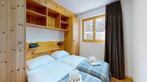 A room with two beds, wooden headboards, a window with curtains, and a view of buildings outside. - Modern apartment with balcony (Haute-Nendaz)